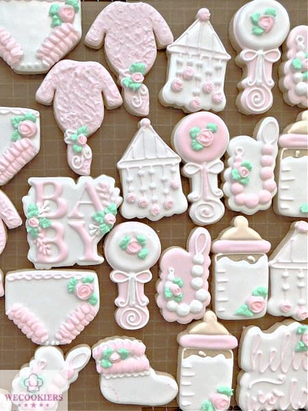 This image reflects soft pink and white baby cookies. It shows an array of shapes: the baby word, knotted nightshirt, mobile, and pacifier clip among others. These pastel pink and white baby girl shower cookies were the perfect set to welcome the little miss. See more inspo, techniques, popular cutters, and RI transfer templates here!
