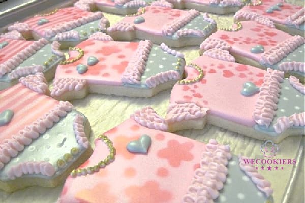 A photo of a group of baby onesies airbrushed with different stencils. Added ruffles, needed necklaces, RI transfer hearts, and a wet-on-wet technique added. Adorable baby girl shower cookies can be achieved through airbrushing patterns. Add extra piping techniques for ruffles and necklaces. More ideas, popular cutters, & RI templates for babies here: