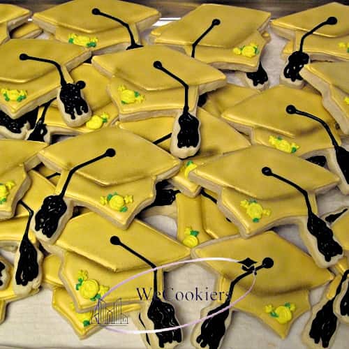 The image reflects graduation sugar cookies in the shape of a cap. They are beautiful yellow with shadows of gold and a yellow rose with green leaf to top it off.